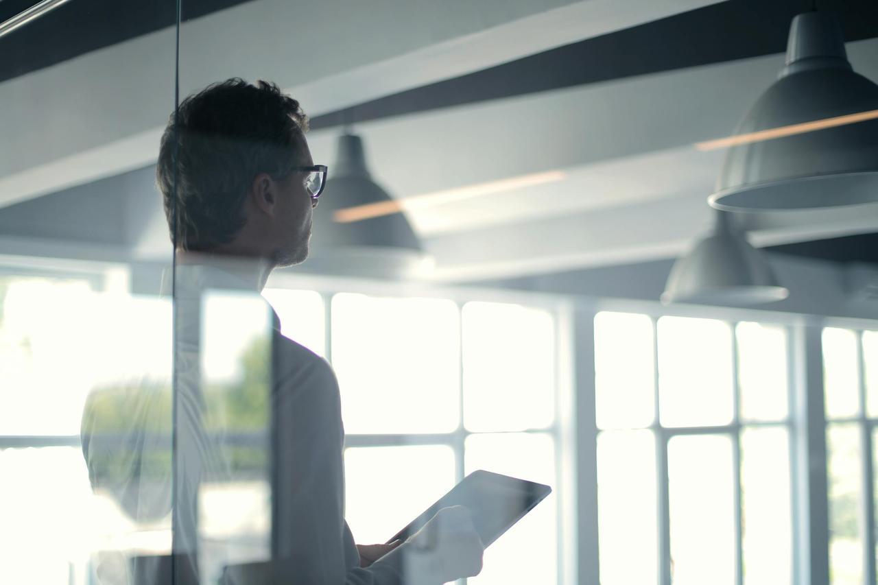 A businessman holding a tablet in an office, looking thoughtfully through a glass wall.
