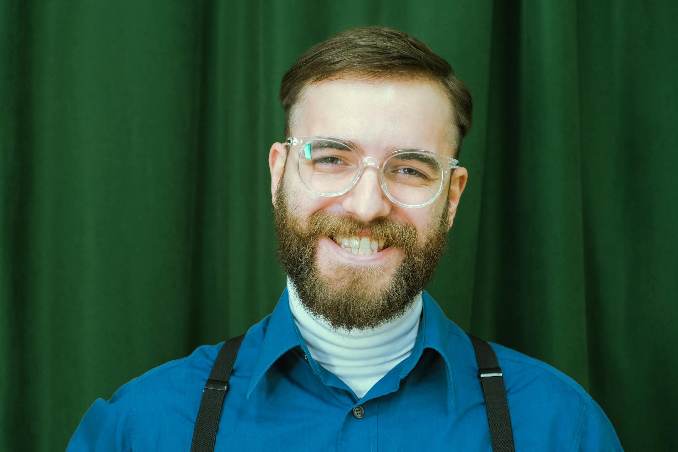 Portrait of a smiling man with beard and eyeglasses against a green curtain background.