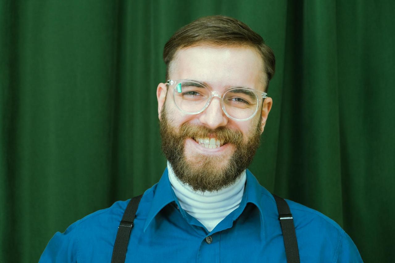 Portrait of a smiling man with beard and eyeglasses against a green curtain background.