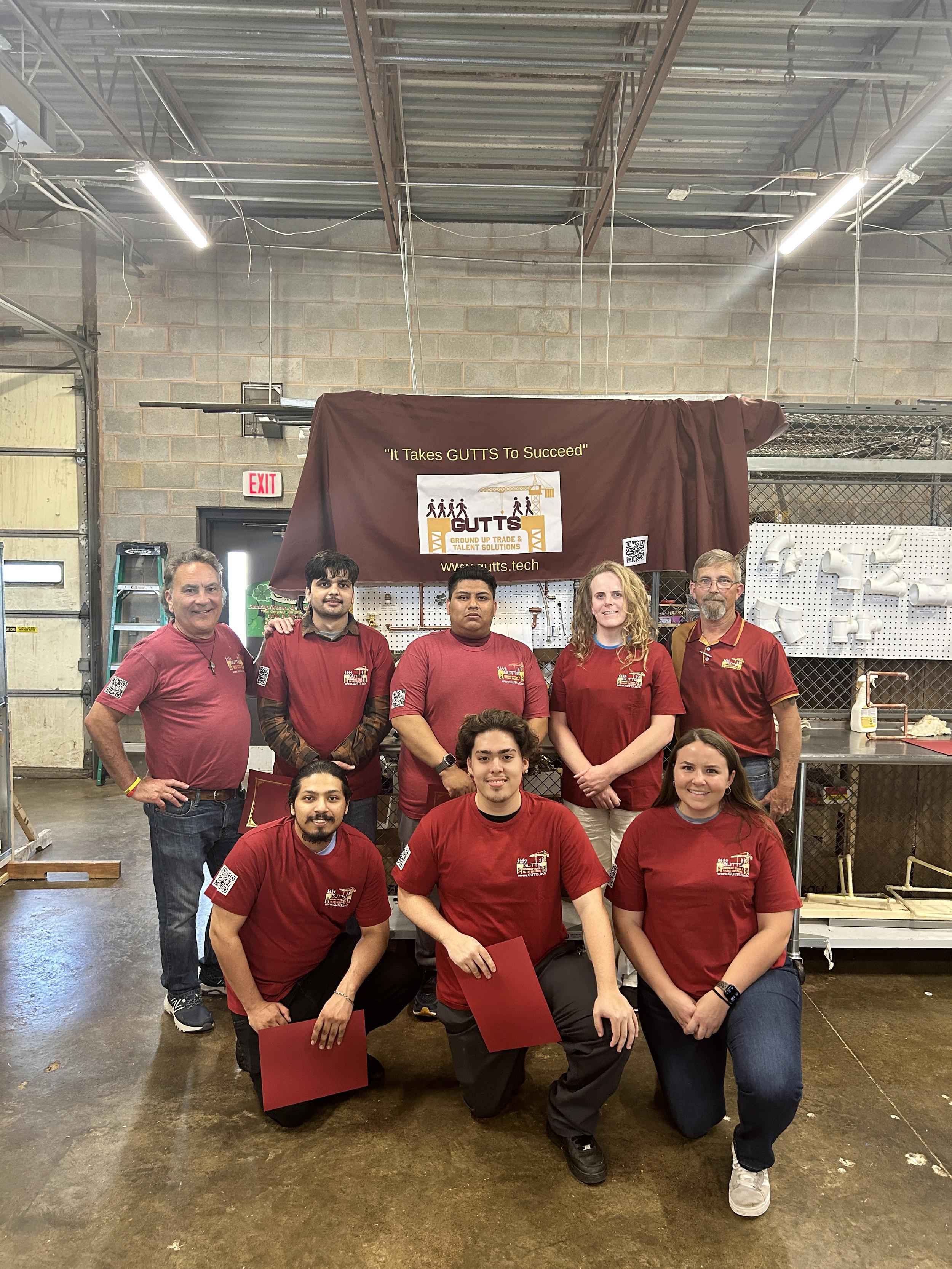 Group of GUTTS Plumbing & HVAC Program graduates standing together on graudation day in front of GUTTS Signage.