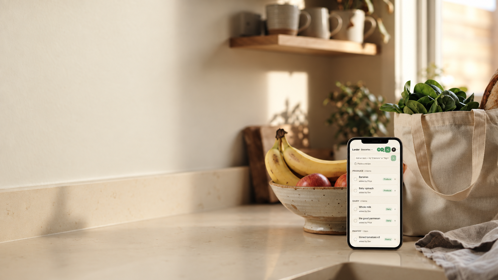 A phone propped against a fruit bowl on a sunlit kitchen counter shows the Larder grocery list, a canvas tote of spinach beside it