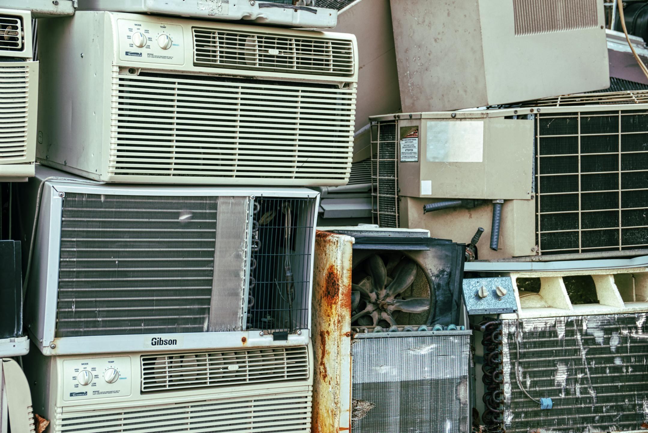 A collection of broken and rusted air conditioners piled in an outdoor junkyard.