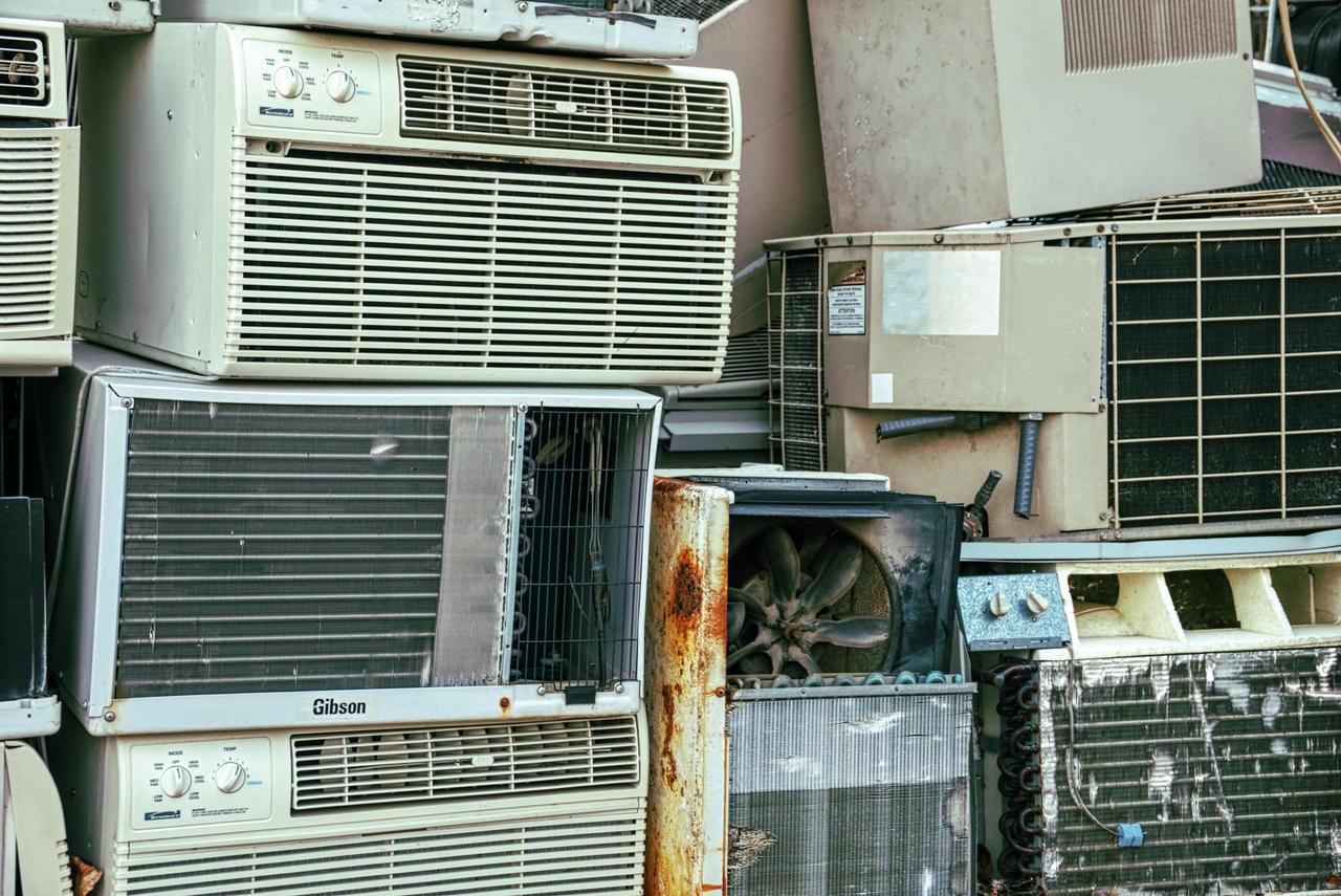A collection of broken and rusted air conditioners piled in an outdoor junkyard.