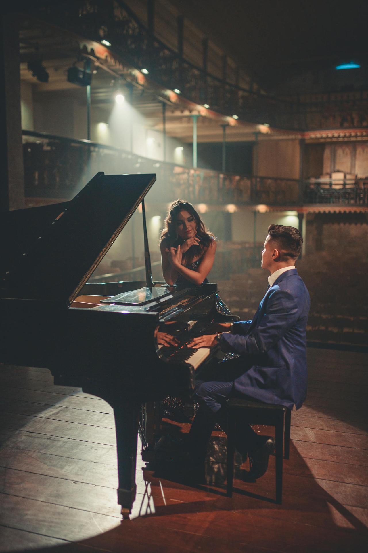 A man playing the piano with a woman nearby in an elegant concert hall setting.