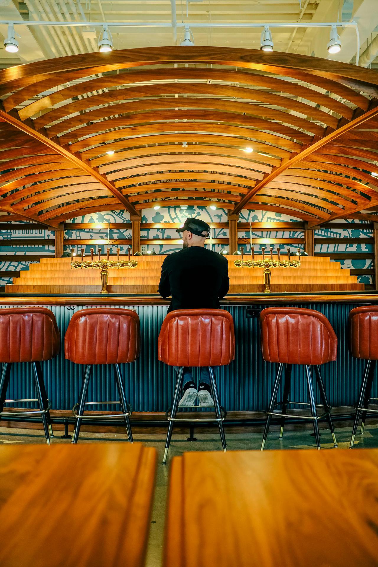 A man sits alone at a bar with a unique wooden arch ceiling, exuding a vintage feel.
