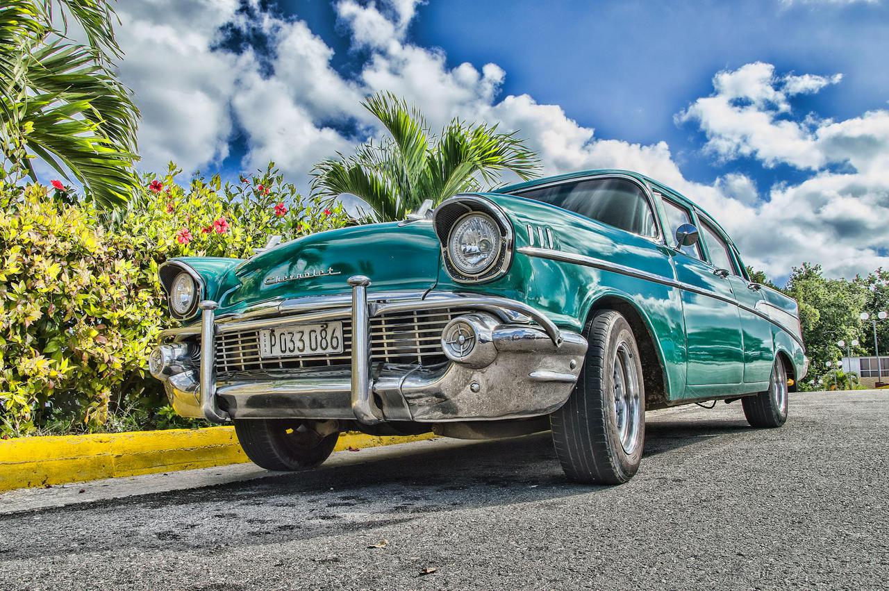 A beautifully restored classic car on a sunny road in Havana, Cuba, with lush foliage and a vibrant sky.