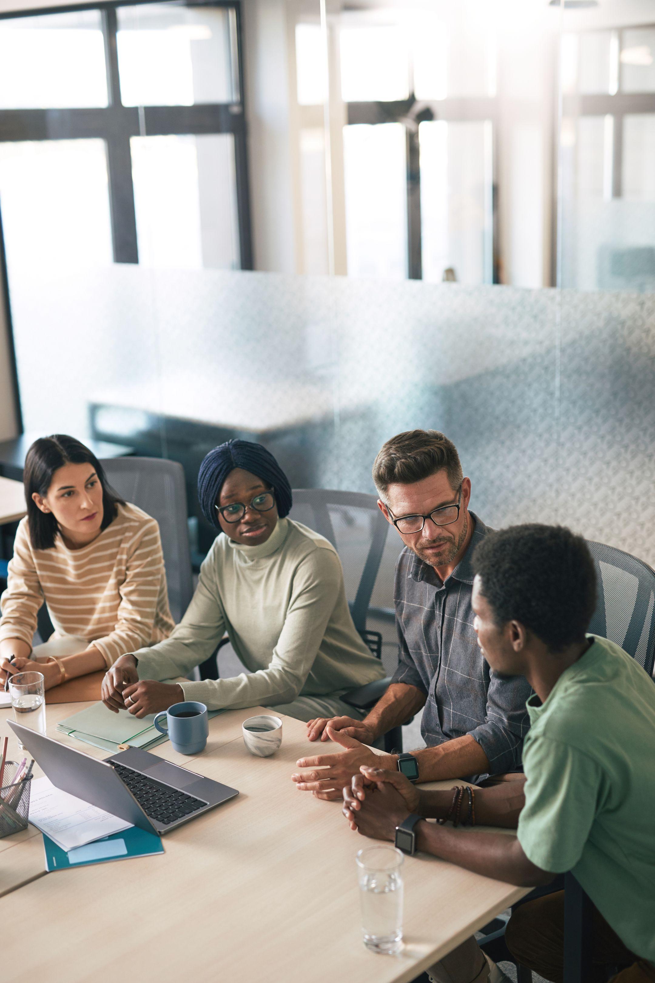 A diverse group of colleagues having a discussion in a modern office setting with laptops and coffee cups.