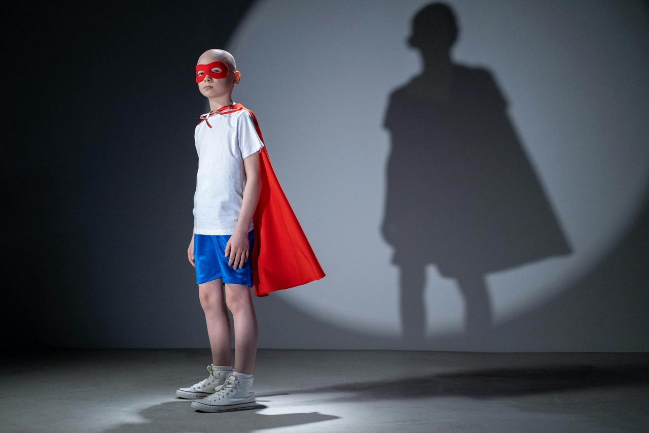 Child in a superhero costume poses confidently with a shadow on a studio wall, symbolizing strength and dreams.
