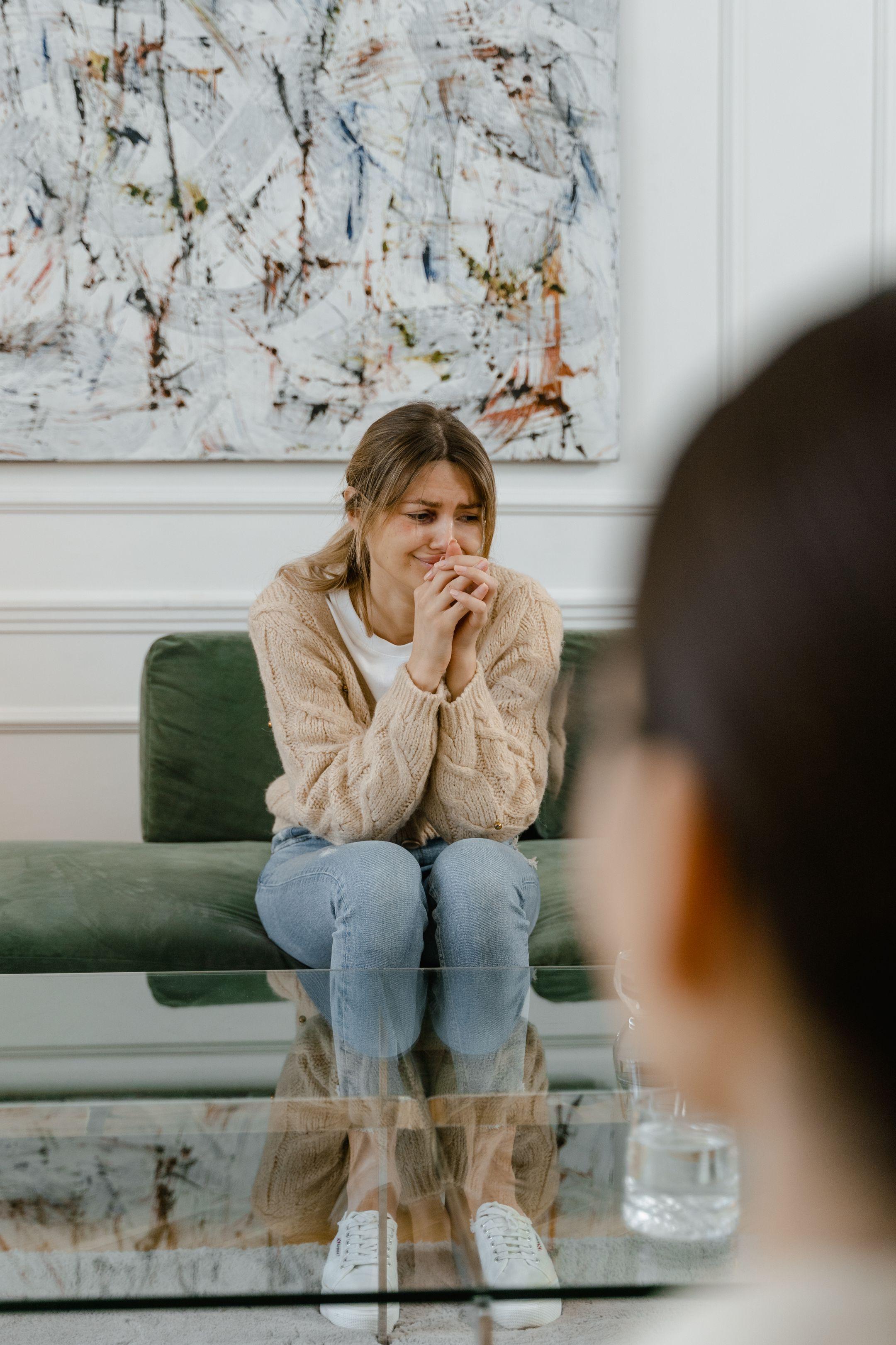 A woman sits on a sofa visibly distressed during a therapy session, hands clasped.