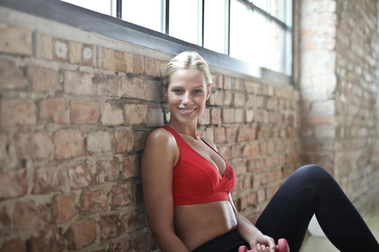 A fit woman in a red sports bra smiling while sitting against a brick wall after a workout.