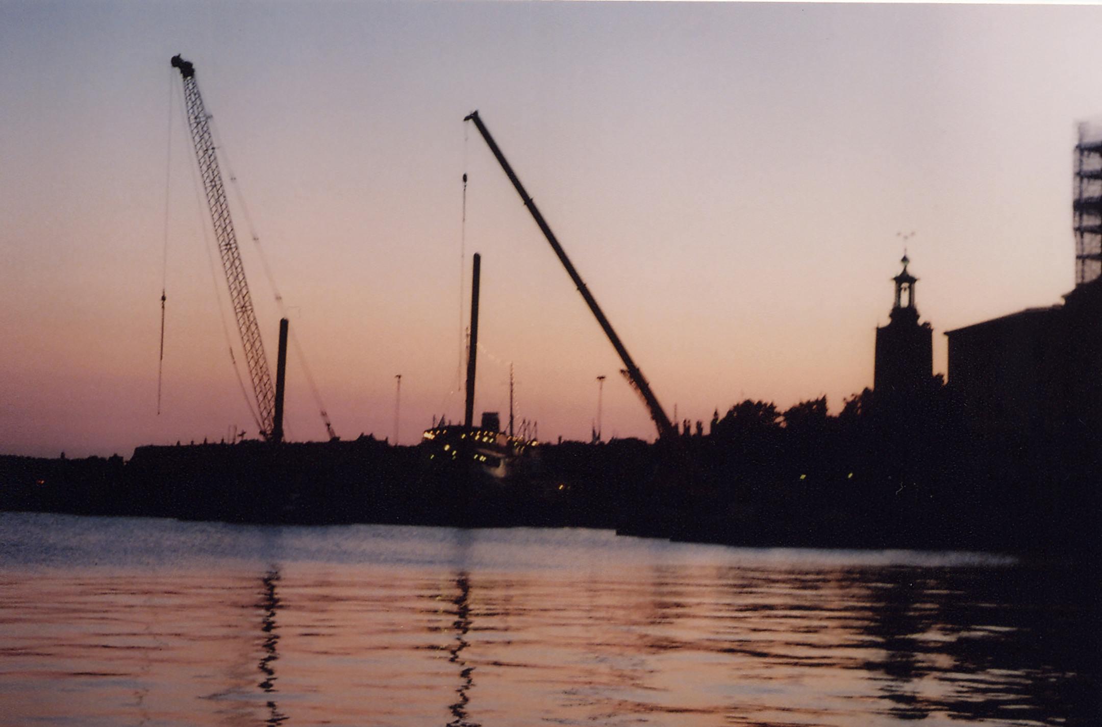 A scenic view of a construction site and cranes silhouetted against a sunset sky by the river.
