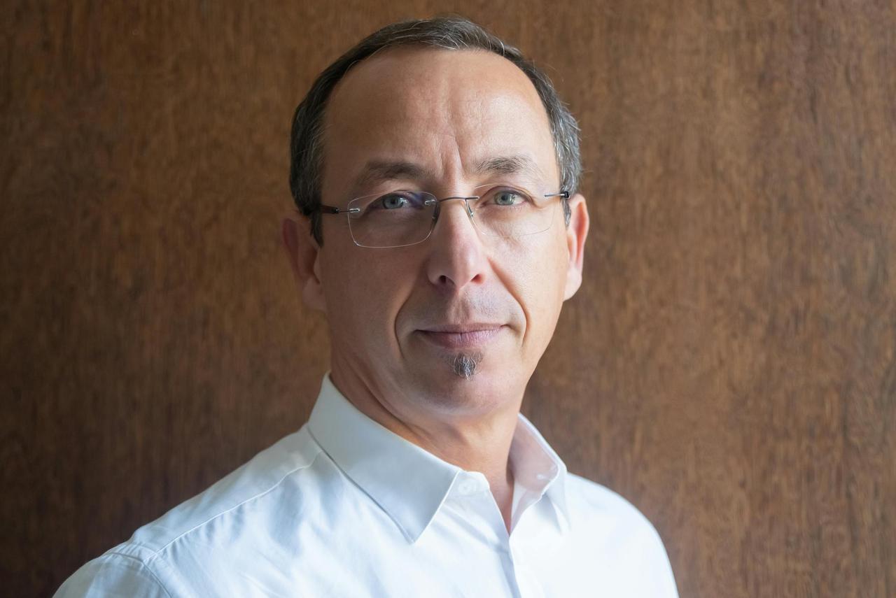 Close-up portrait of a middle-aged man wearing eyeglasses, dressed in a white shirt, set against a wooden background.