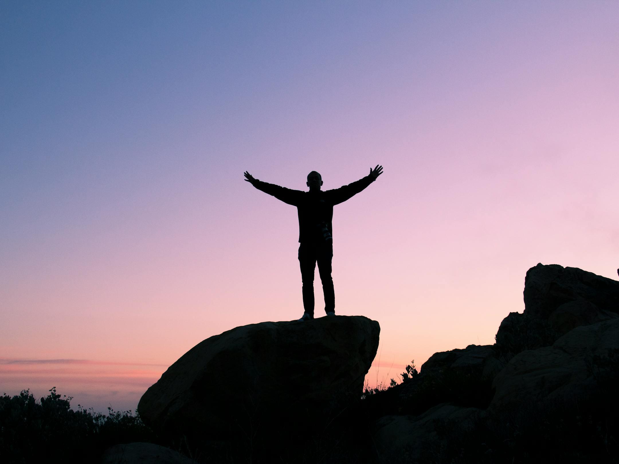 Silhouette of a man on a rock with arms raised, embracing the sunset sky.