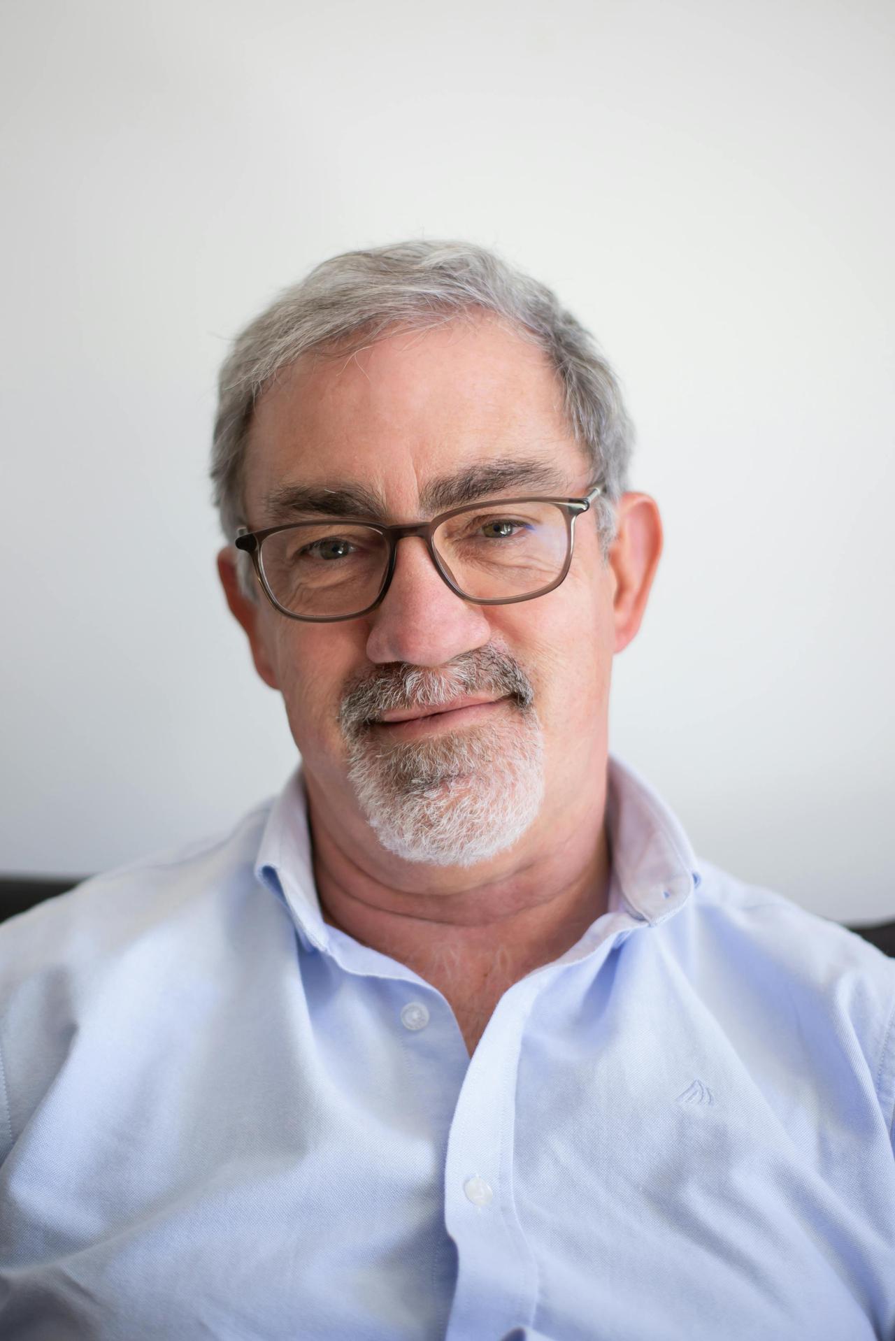 Close-up portrait of a smiling mature man with glasses and gray hair, wearing a blue shirt.