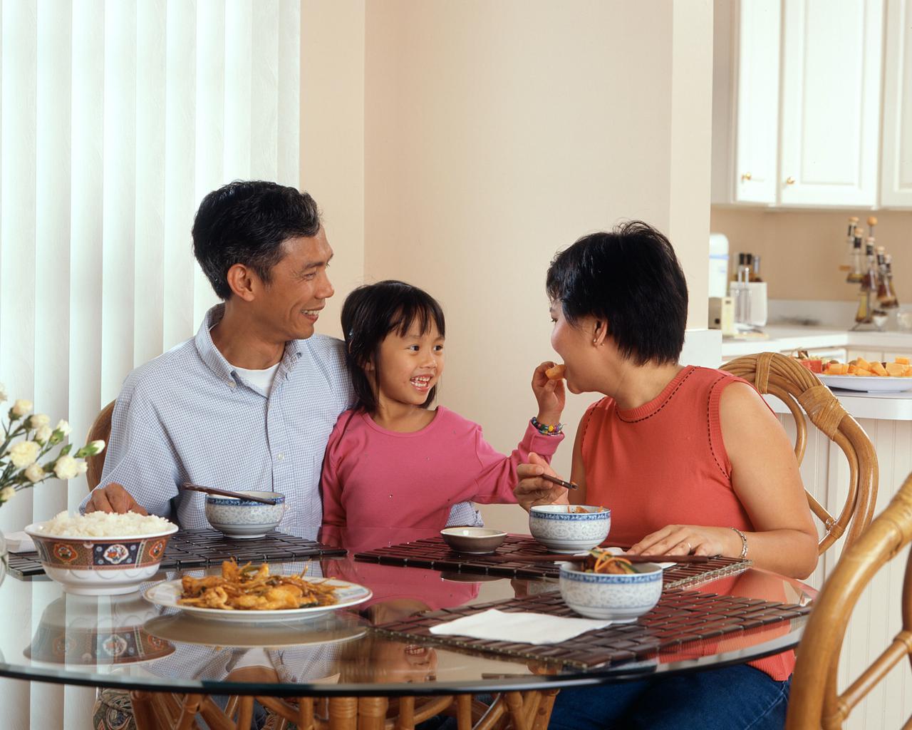 An Asian family, an adult male and female are seated around a table eating a meal with a young female standing in between the adults.