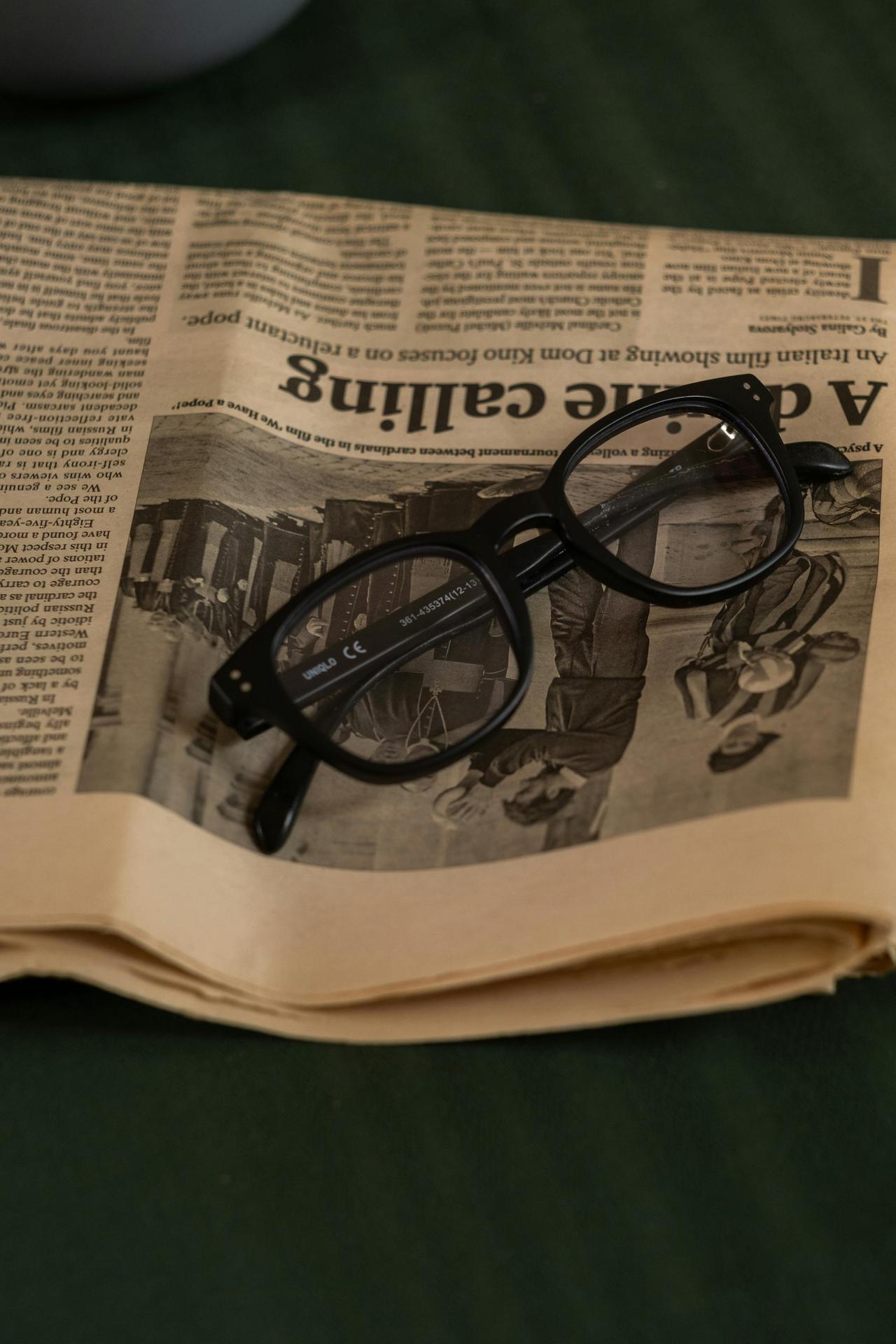 Black rimmed glasses resting on top of a newspaper on a green surface.