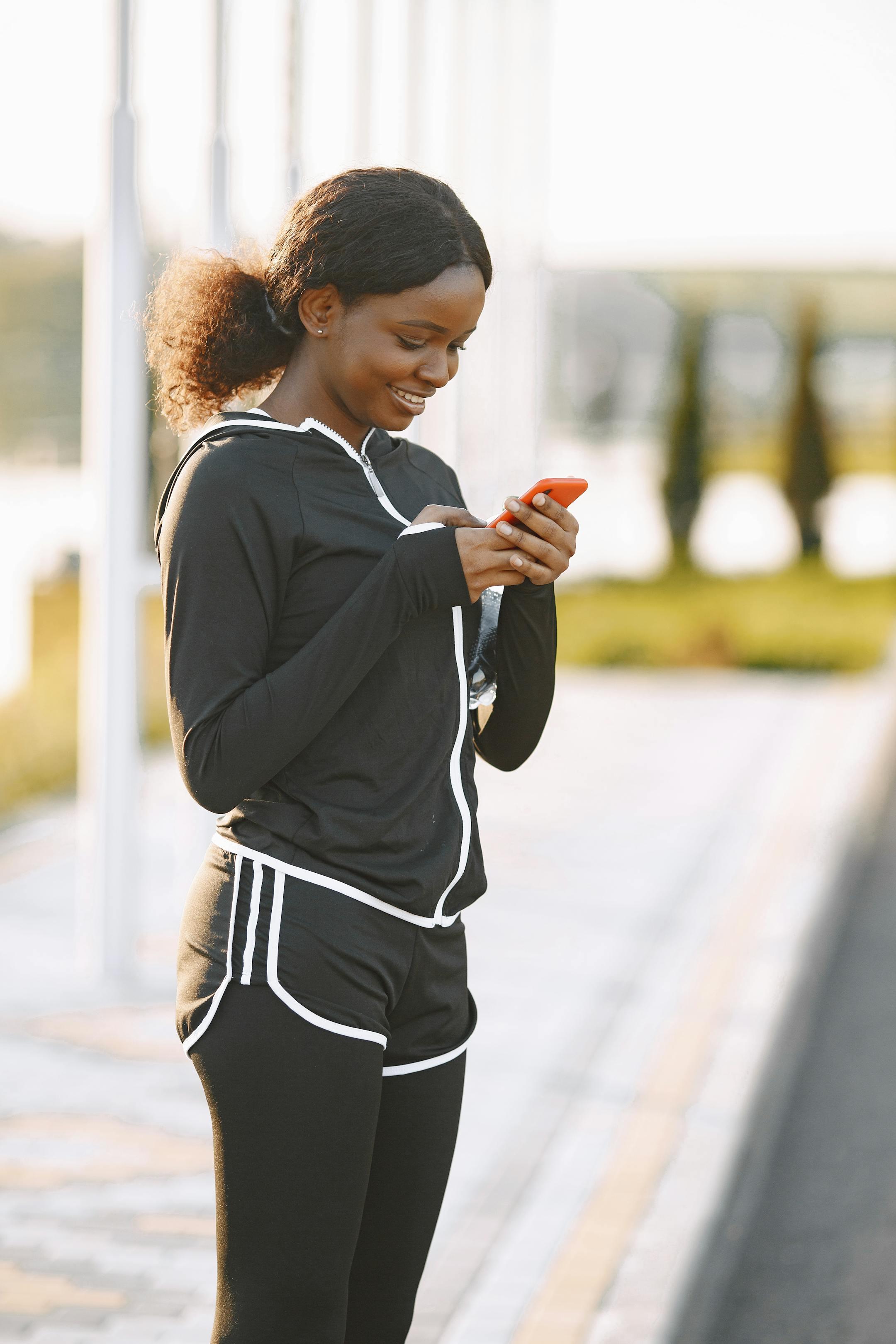 A woman in activewear smiling while texting on her phone outdoors, enjoying a sunny day.