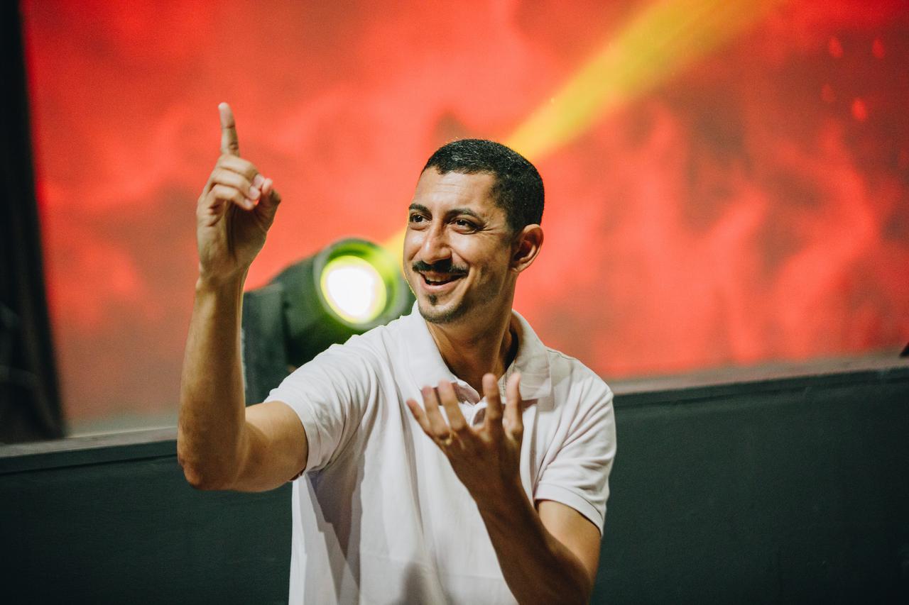 A man with a mustache and white shirt on a stage with a red backdrop using sign language. An ASL interpreter in Miami is indispensable when dealing with people who are hearing impaired.