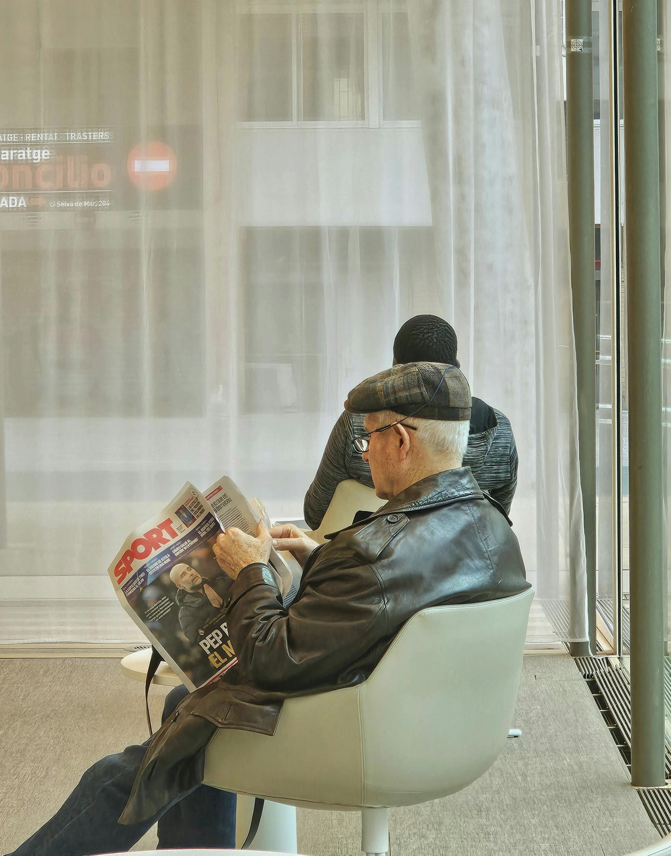 Elderly man reading a sports newspaper in a cozy indoor setting, seated in a modern chair.