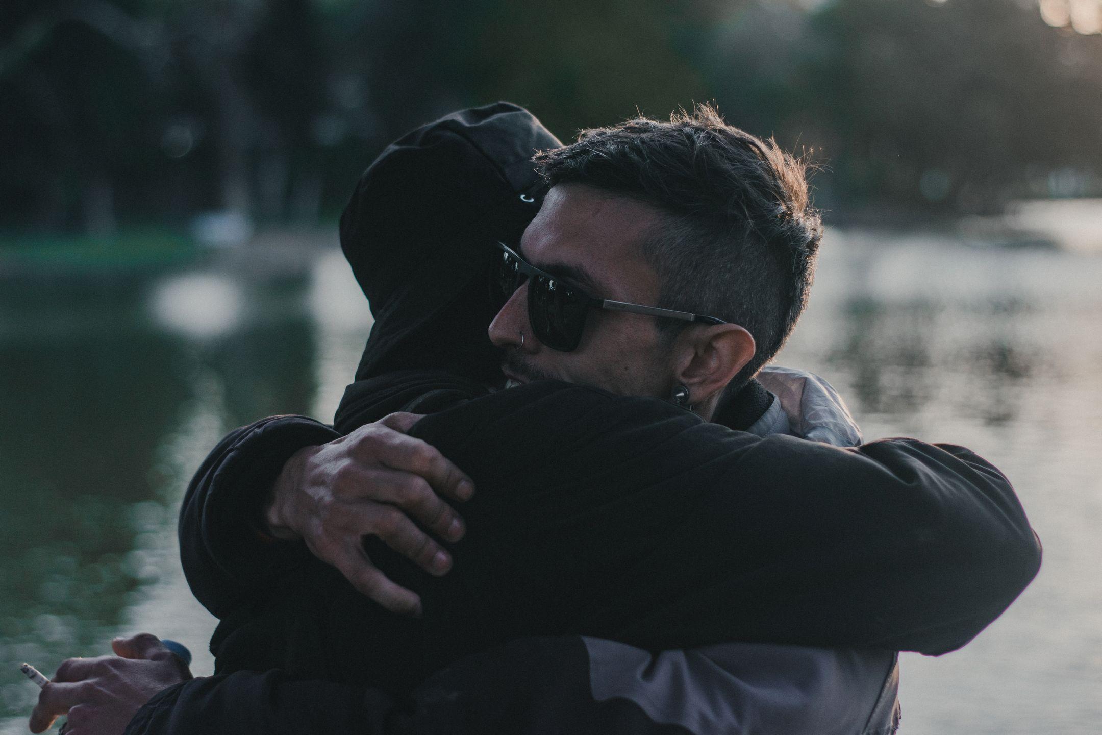 A heartfelt embrace between two adults near a calm lakeside during daylight.