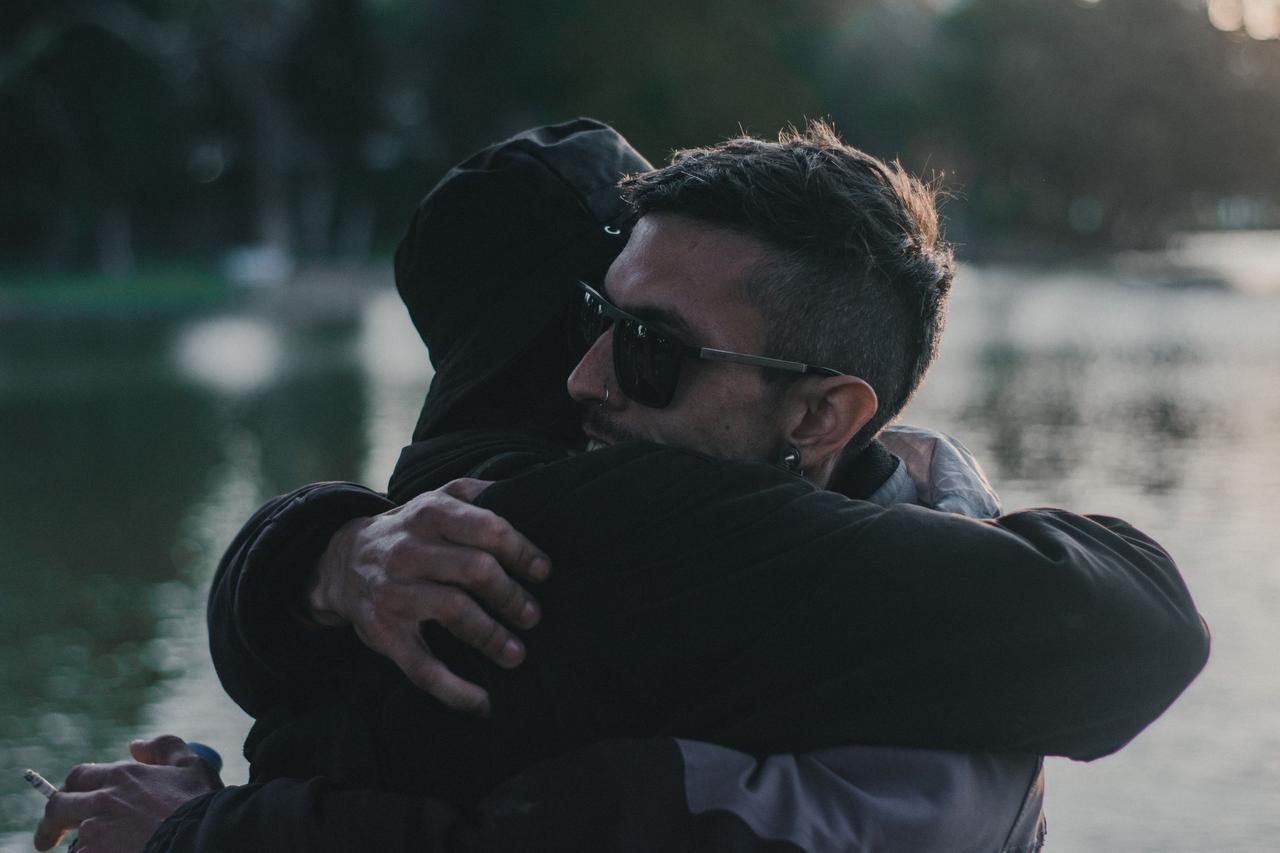A heartfelt embrace between two adults near a calm lakeside during daylight.