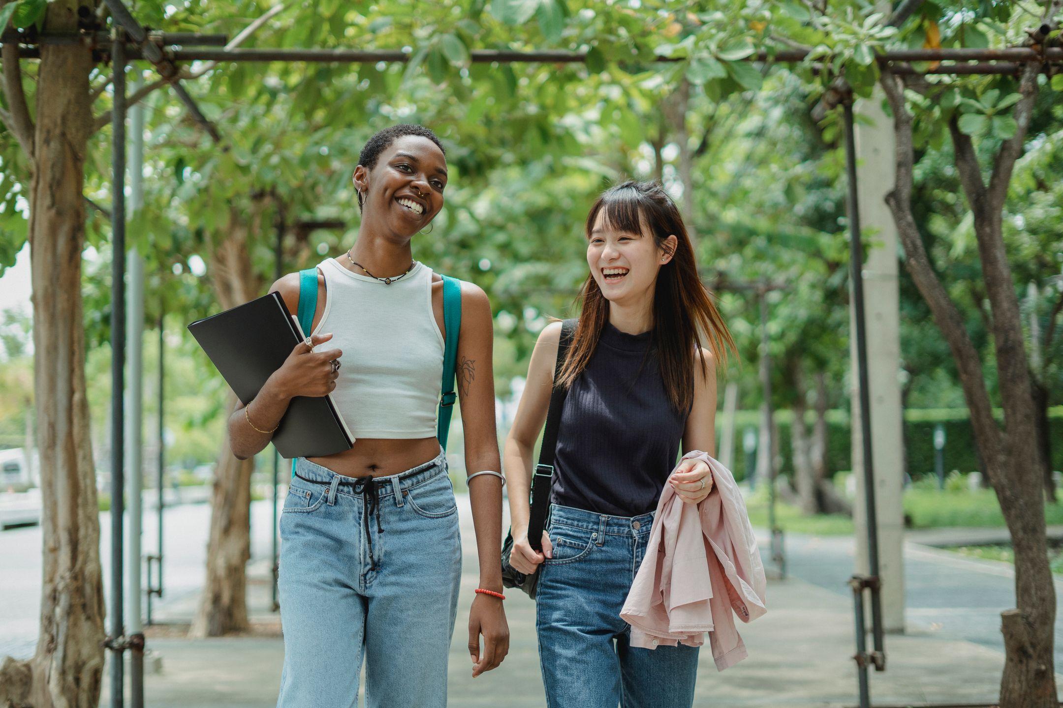 Positive young multiracial female friends in casual outfits with backpacks having conversation while walking along alley