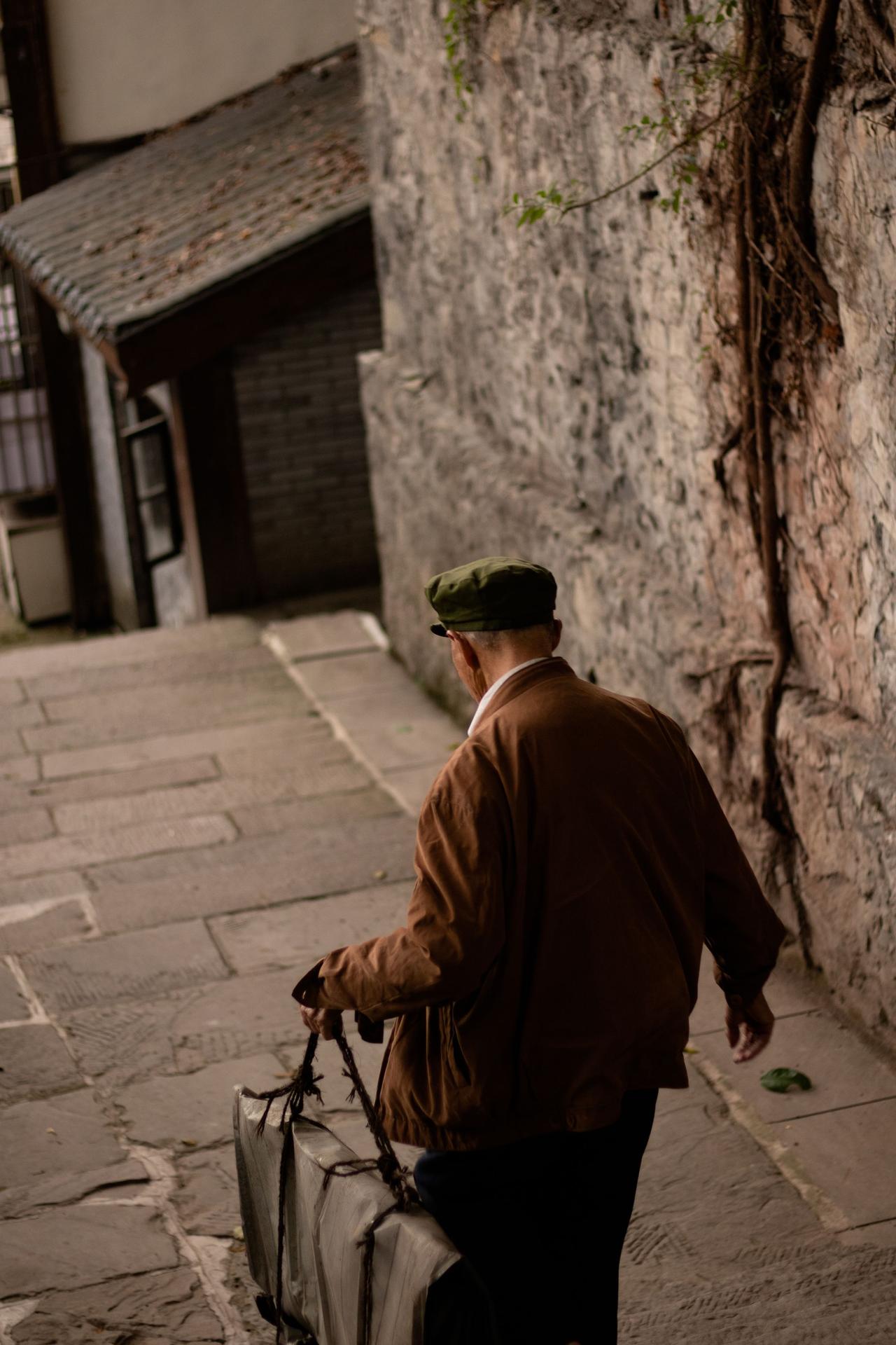 A man carrying suitcases walks down an old stone alleyway with historical buildings.