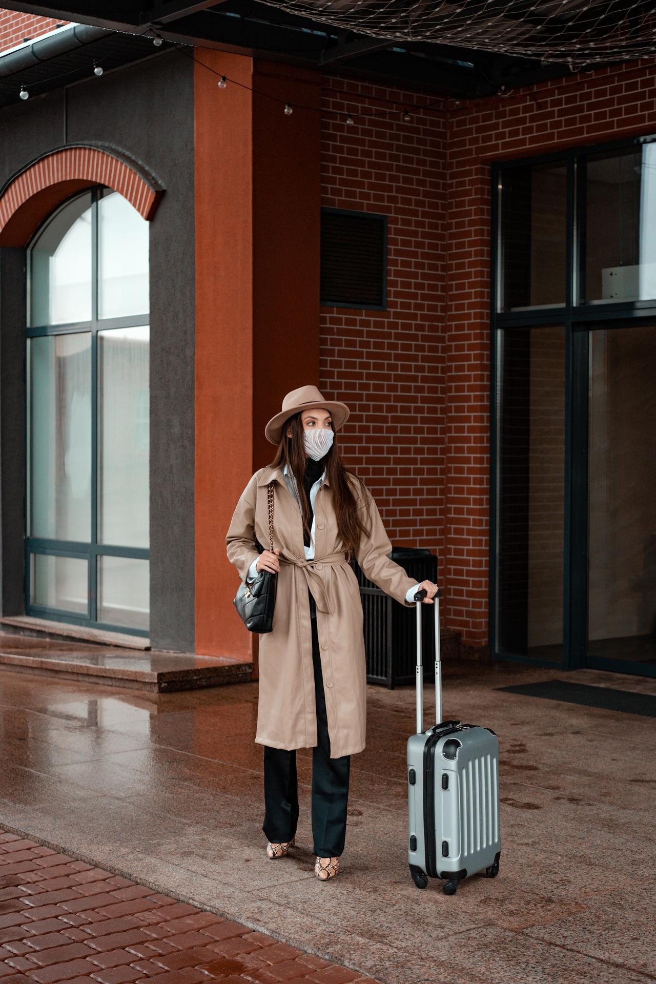 Stylish woman in coat and hat with suitcase outdoors, showcasing travel fashion.