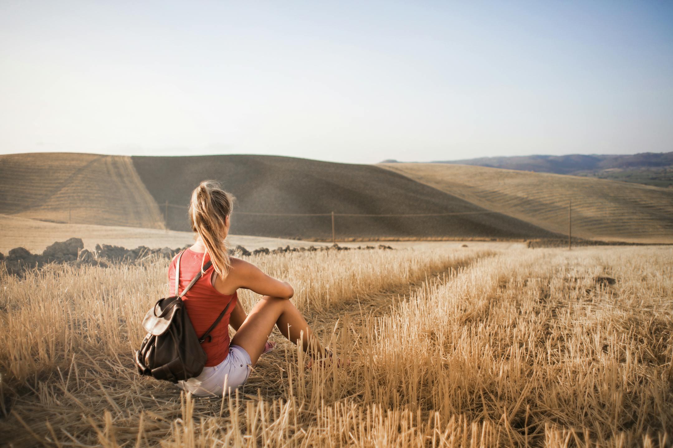 Woman sitting with backpack in rural wheat field, enjoying serene summer landscape.