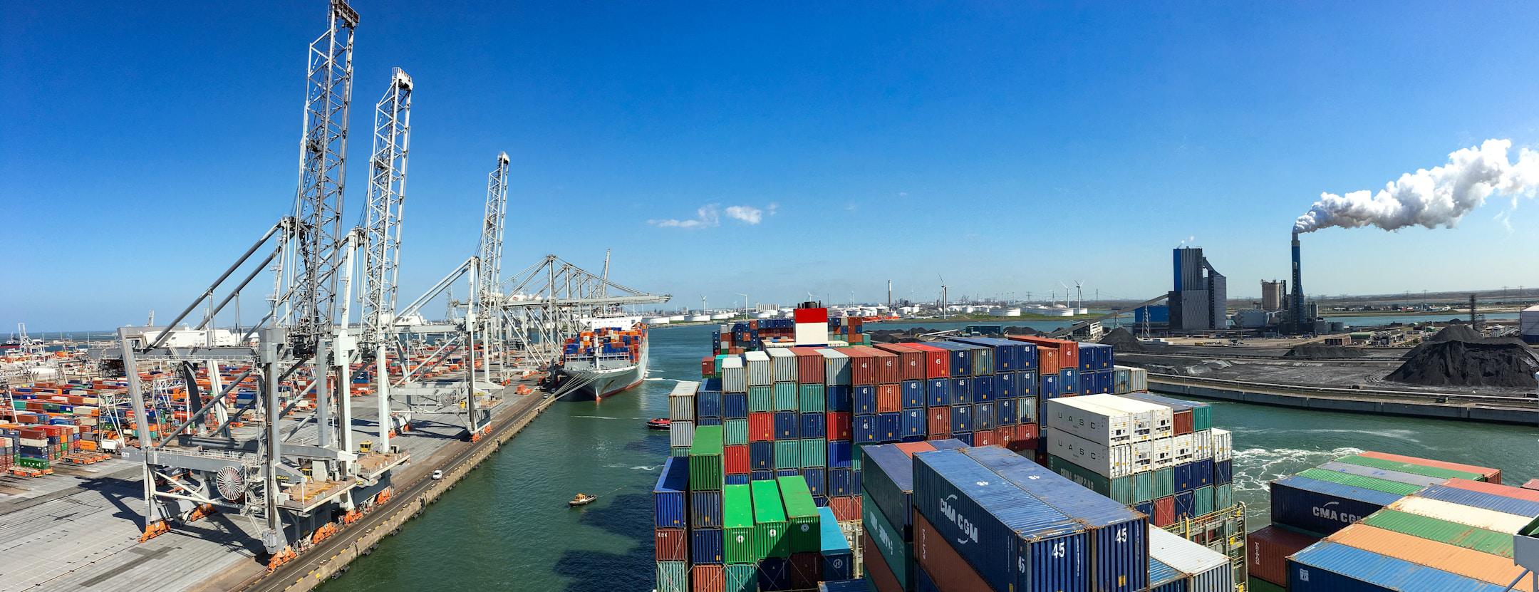 A panoramic view of a busy shipping port featuring rows of colorful shipping containers stacked high along the docks. Large industrial cranes are positioned along the water's edge, loading and unloading cargo from container ships. In the background, an industrial facility with a tall smokestack emitting white smoke contrasts with the clear blue sky. The port is surrounded by water, with small tugboats visible near the ships, highlighting the scale and activity of the maritime industry.