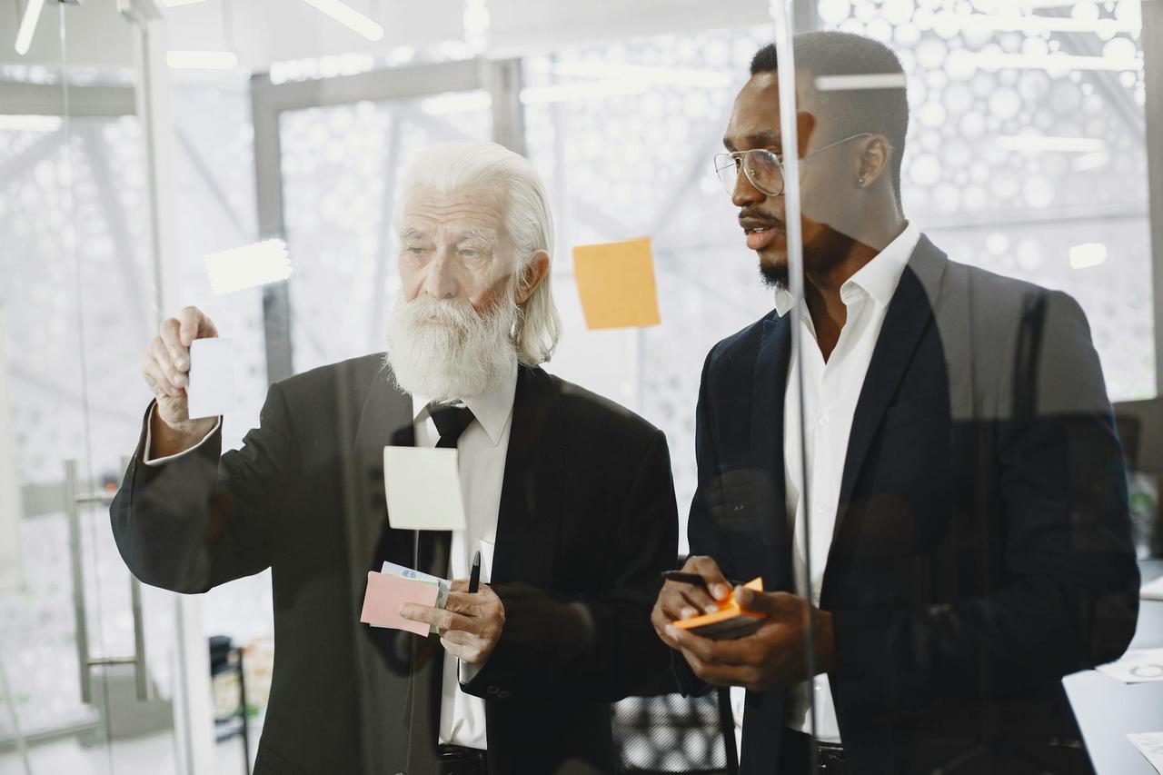 Two men in suits discussing ideas with sticky notes on a glass wall in an office.