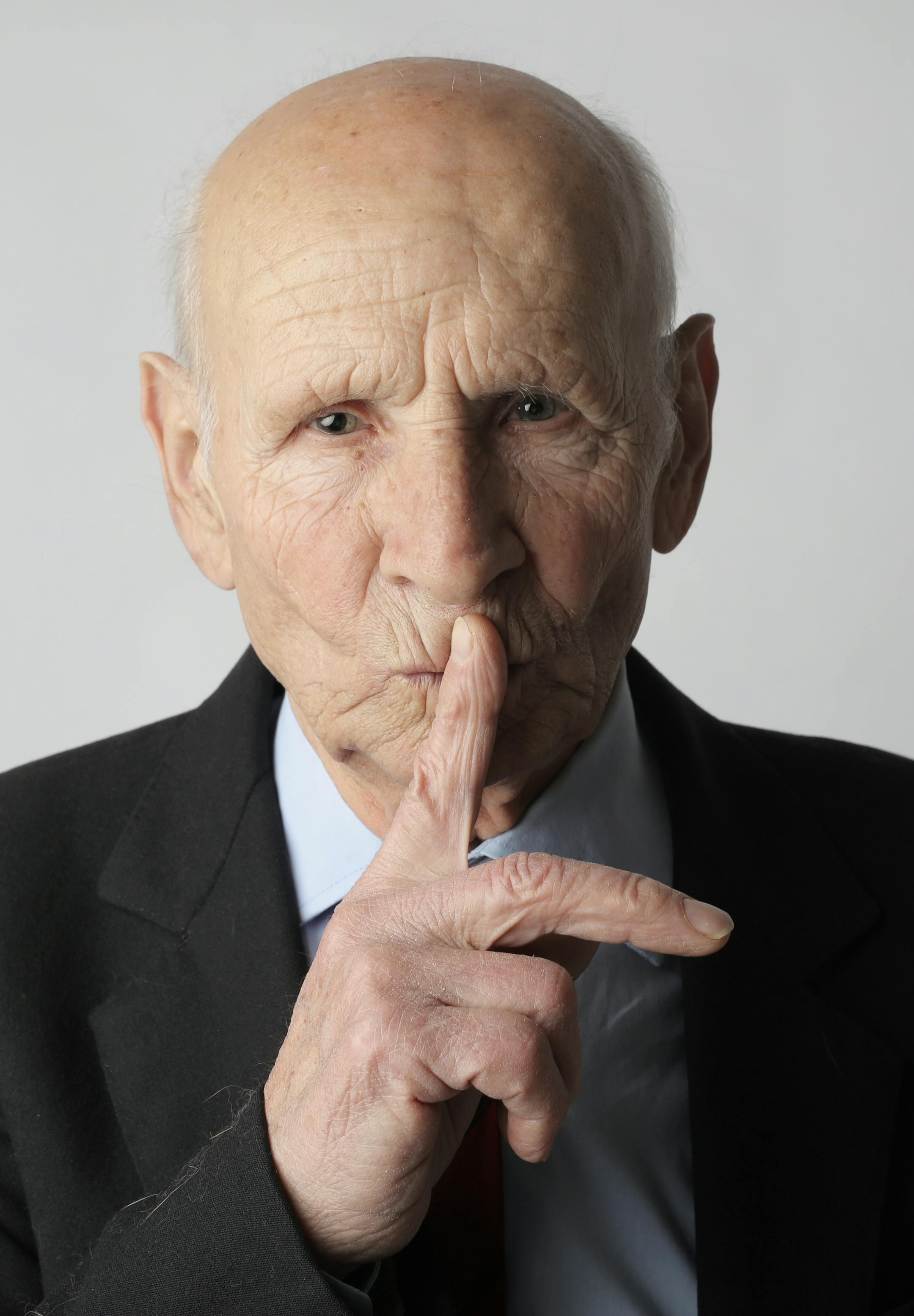 Portrait of a senior man wearing a suit, gesturing for silence in a studio setting.