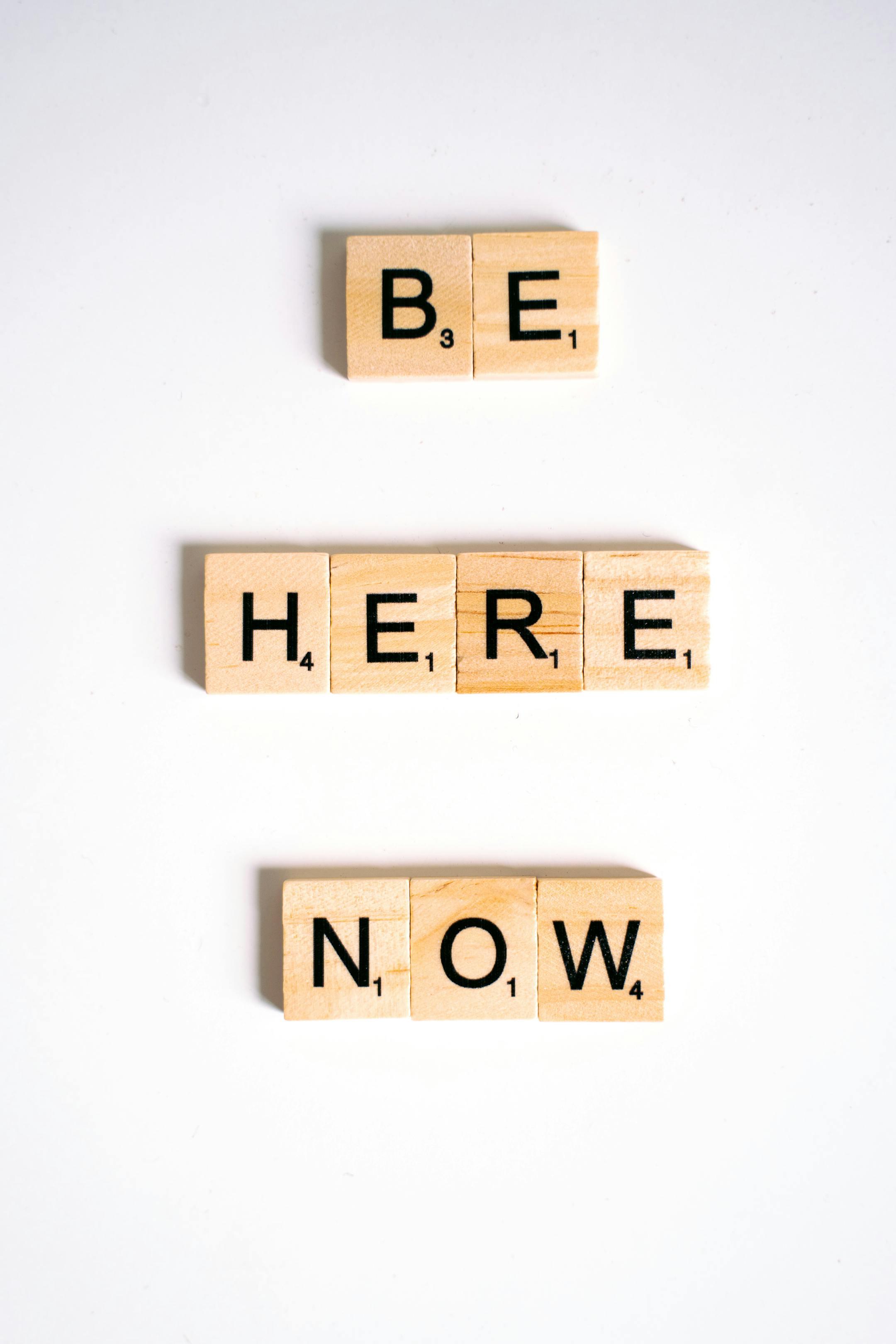 Scrabble tiles spelling 'Be Here Now' against a white background, offering inspiration.