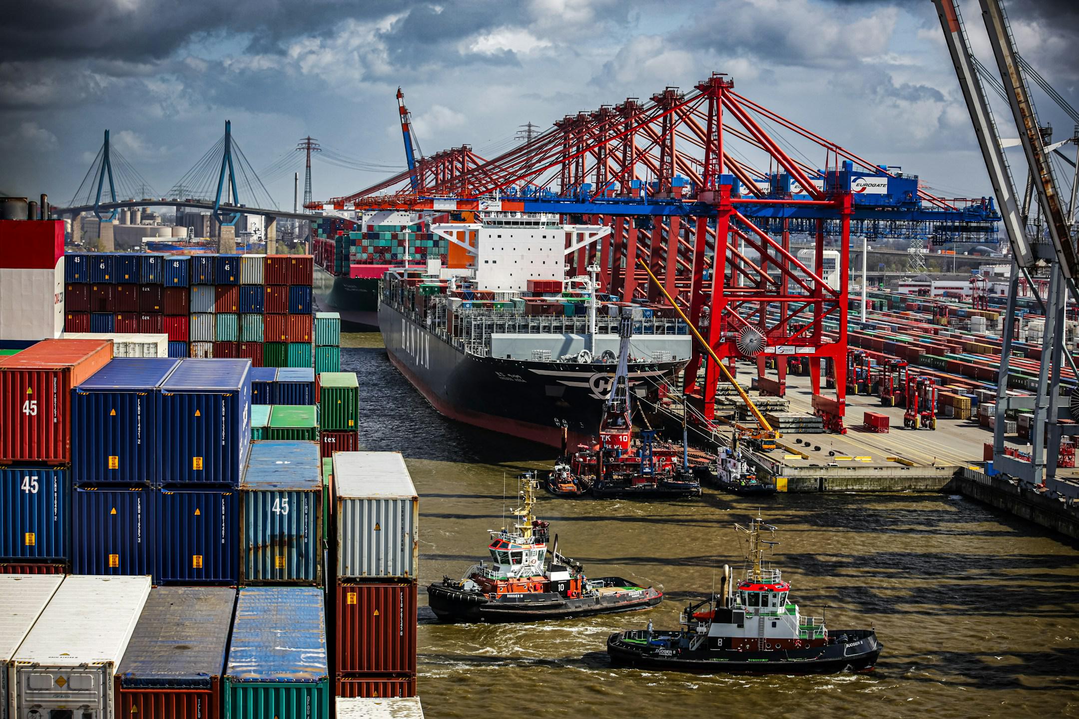 Vibrant port scene with a large cargo ship being guided by tugboats, flanked by towering red cranes and stacks of multicolored shipping containers under a dynamic sky.