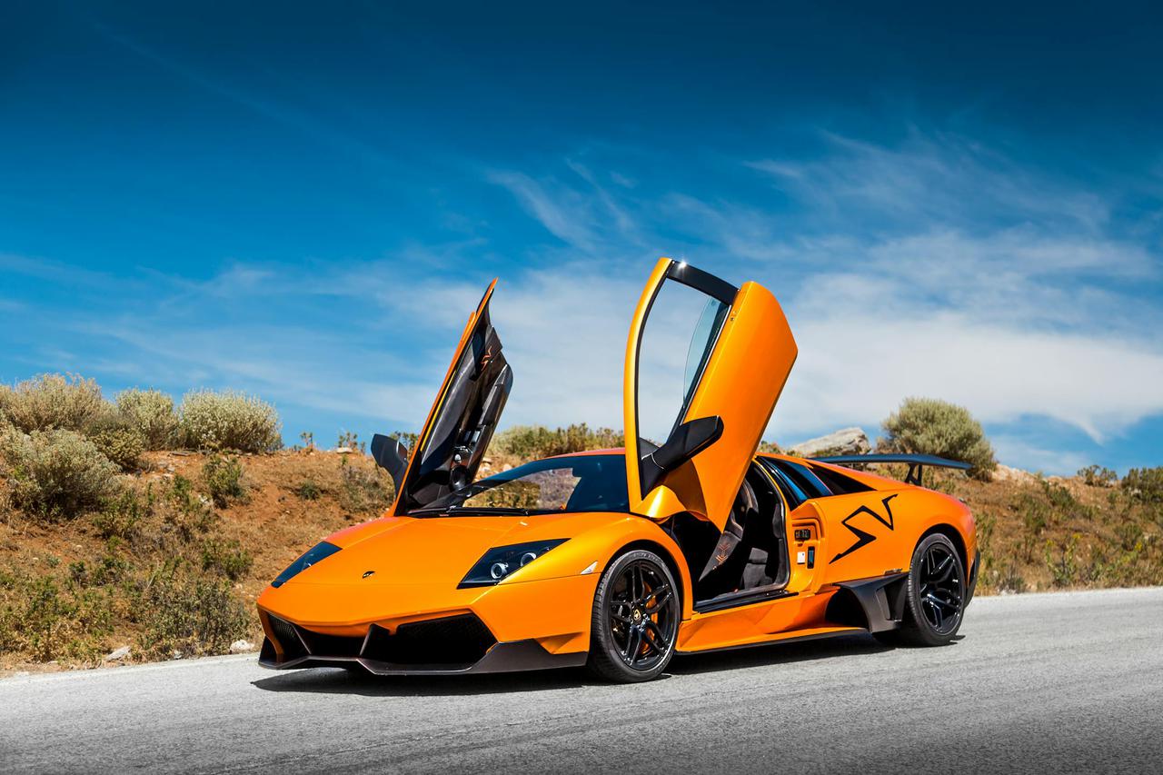 Orange sports car with doors open under bright blue sky on a scenic road.