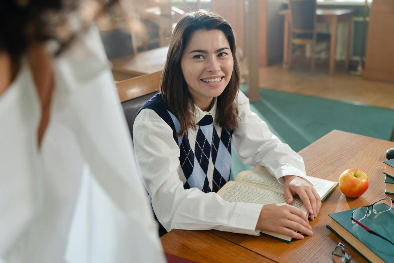 Smiling woman reading a book at a library table, portraying a lively academic atmosphere.