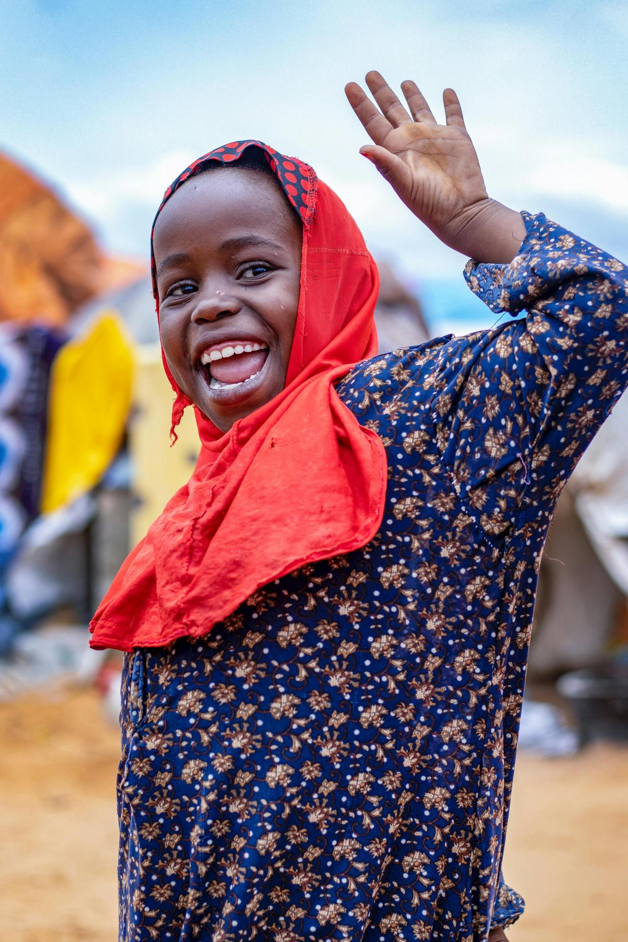 A young girl in a red headscarf smiling and waving outdoors in Galguduud, Somalia.