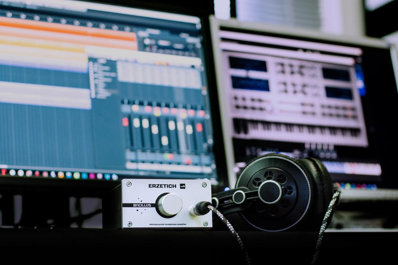 Headphones and headphone amplifier on a desk, monitors with music creation software in the background