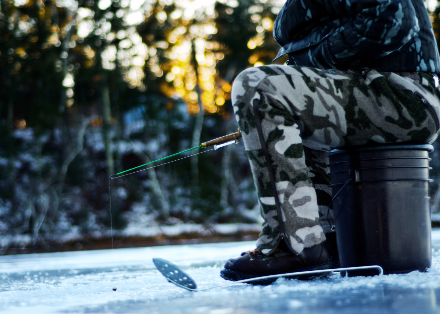 Ice fisherman is sitting on a bucket on top of a frozen lake.  He is holding a custom ice fishing rod over a fishing hole in the ice.