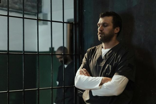 A bearded man in jail uniform stands with arms crossed, reflecting quietly.