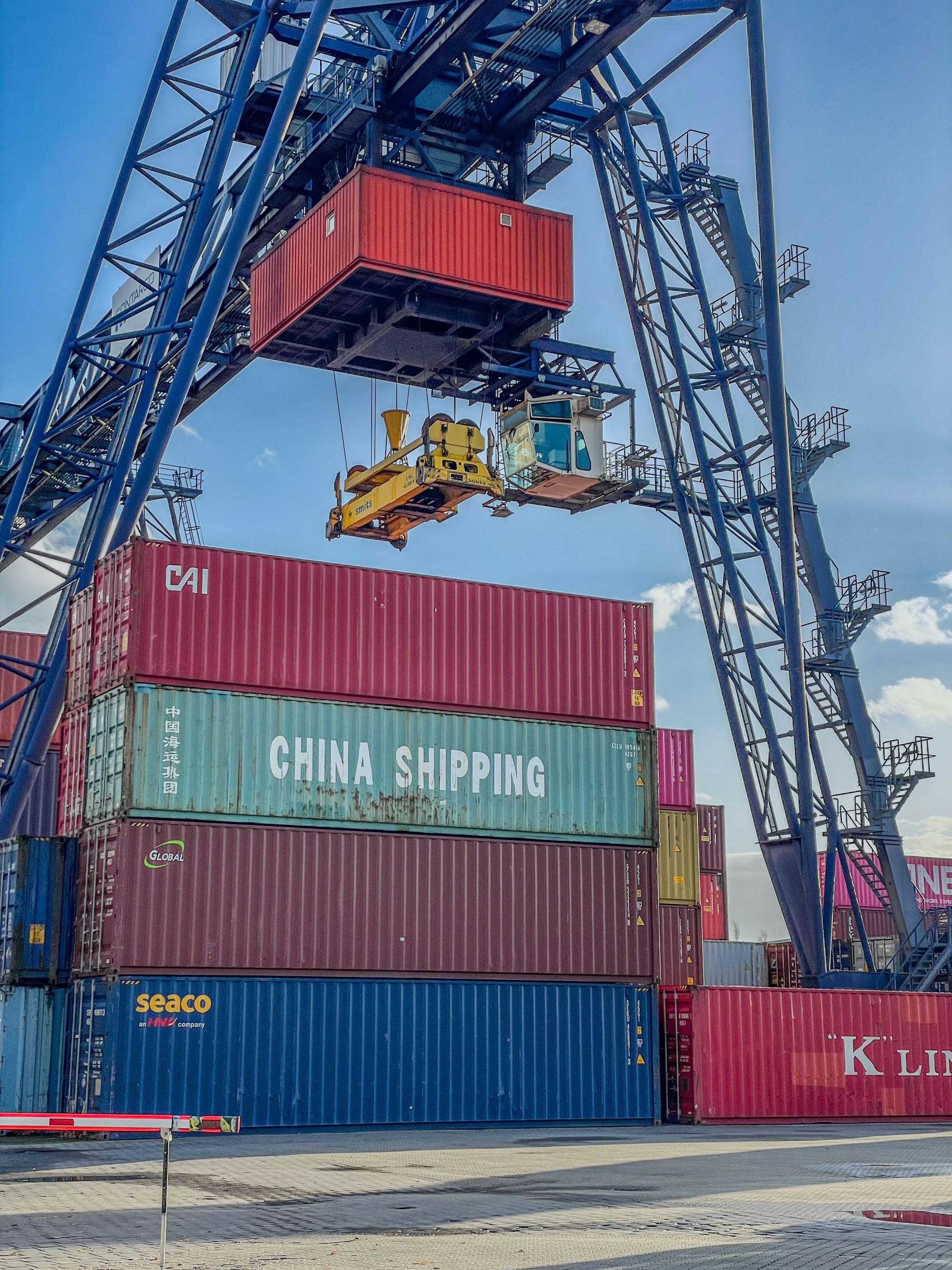 A gantry crane lifting a red shipping container above a stack of various containers with "CHINA SHIPPING" visibly marked, under a blue sky with clouds.