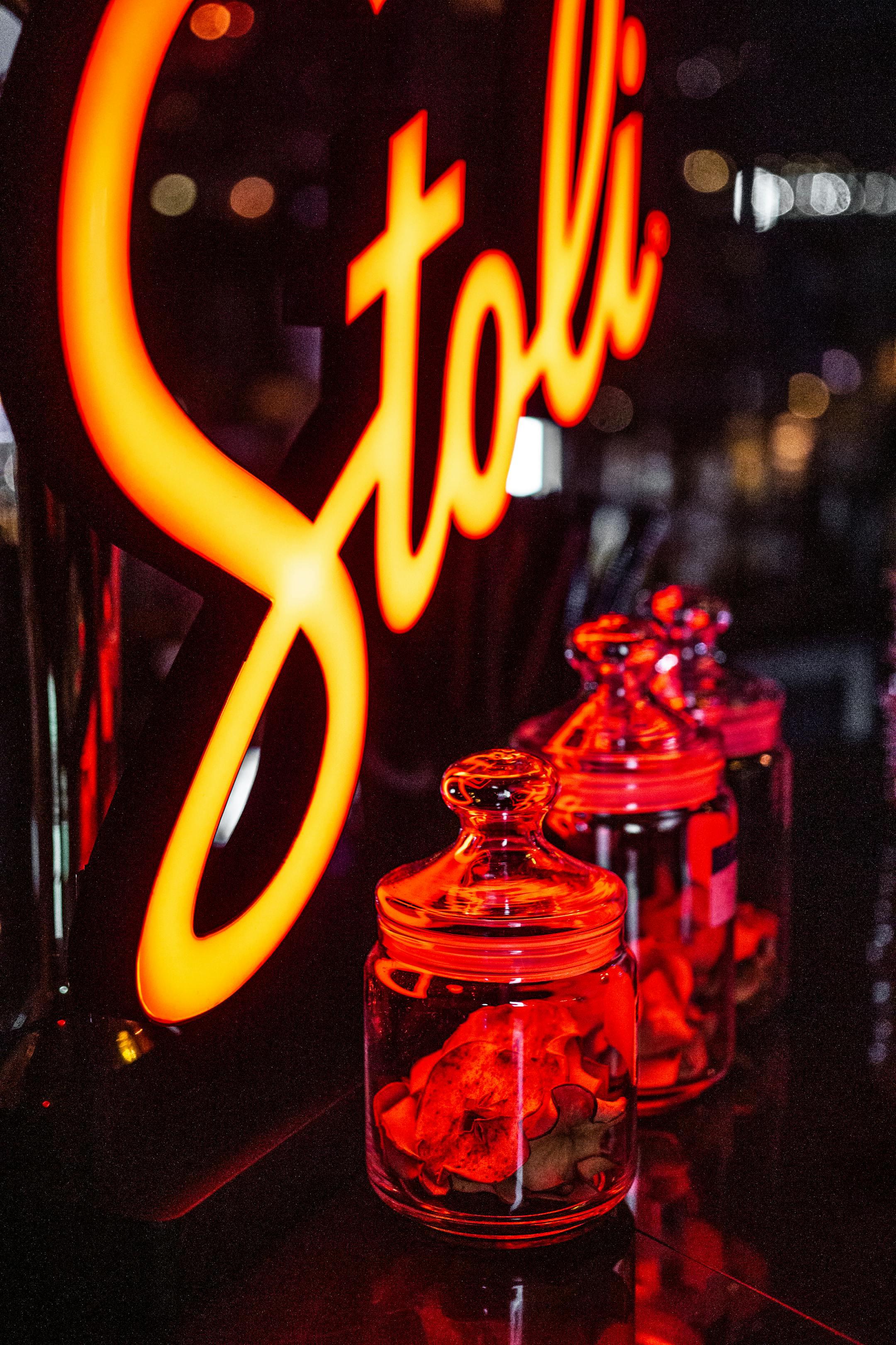 Illuminated red neon 'Stoli' sign with decorative glass jars in a dimly lit bar setting.