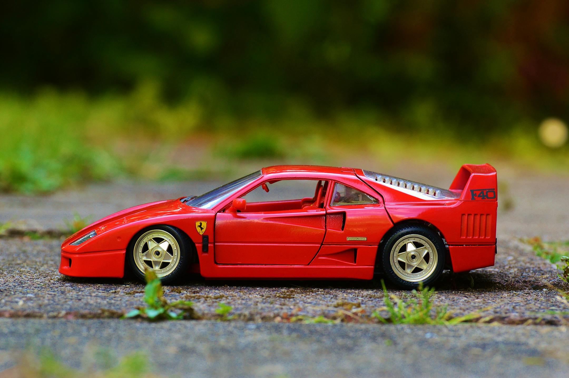 A detailed red Ferrari F40 miniature toy car parked on a sidewalk with greenery backdrop.
