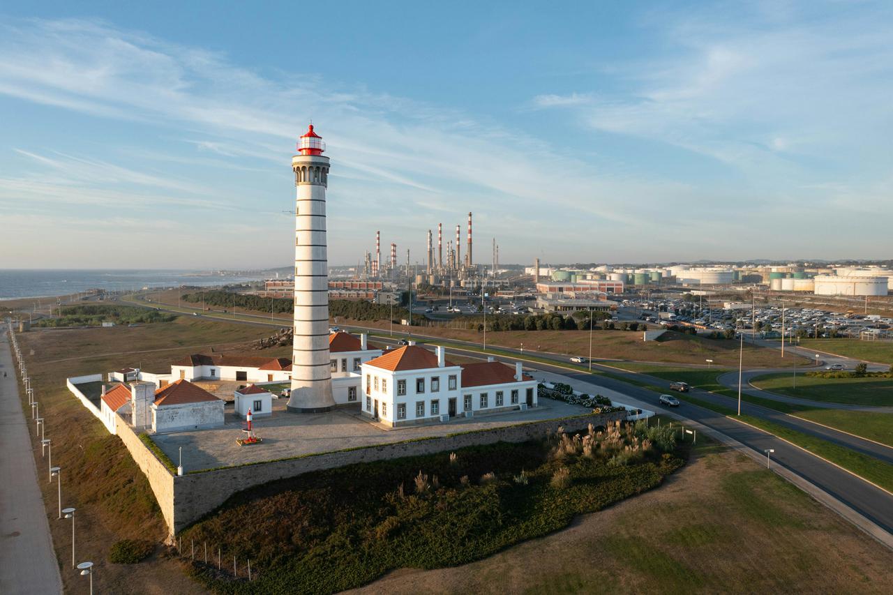 Witness the intersection of past and present in this captivating aerial shot! A classic lighthouse, standing tall with its distinctive spiral pattern, overlooks the vast expanse of the modern industrial complex and the tranquil sea beyond. It's a unique contrast where history meets modernity, guiding the way into the future.