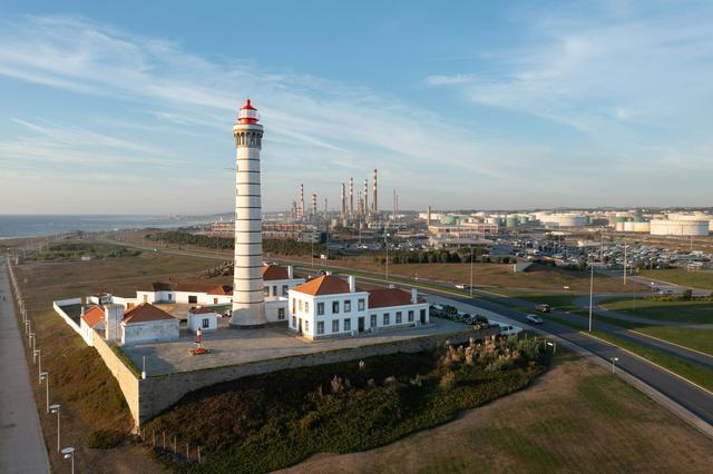 Witness the intersection of past and present in this captivating aerial shot! A classic lighthouse, standing tall with its distinctive spiral pattern, overlooks the vast expanse of the modern industrial complex and the tranquil sea beyond. It's a unique contrast where history meets modernity, guiding the way into the future.