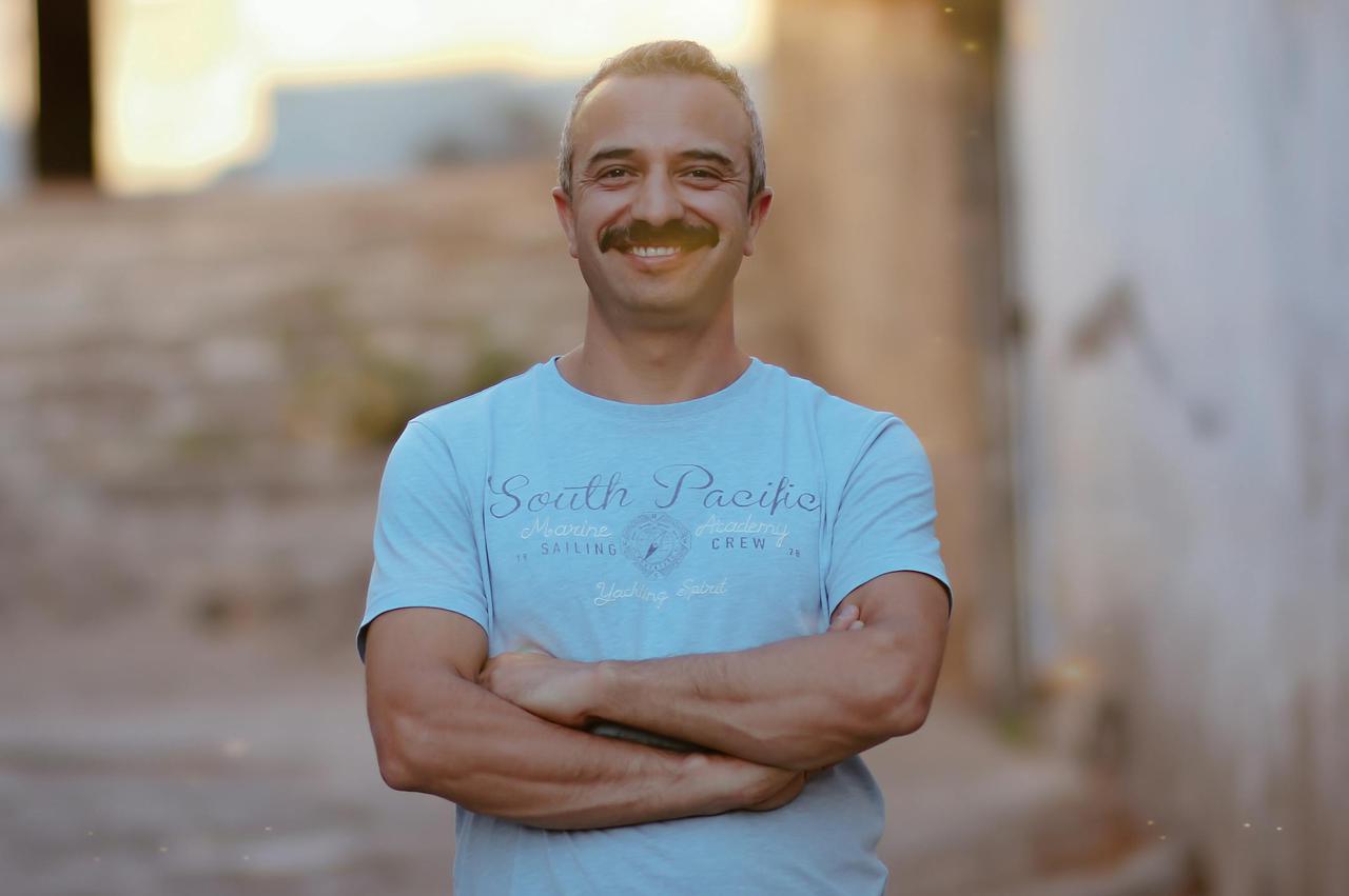 Portrait of a smiling man with a moustache standing on a sunny city street.