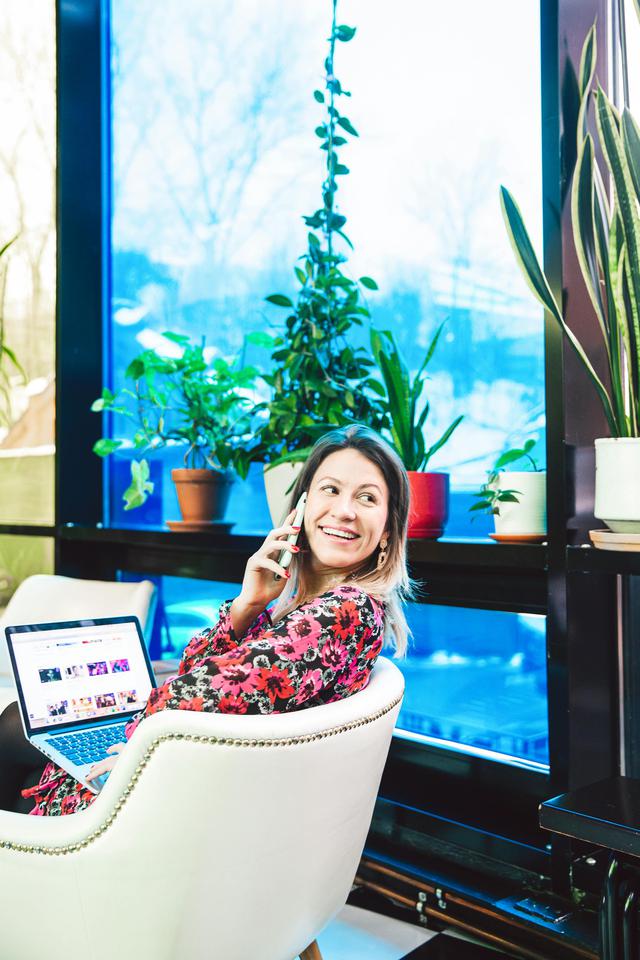 Smiling woman using phone and laptop in an indoor office setting with plants and large windows.