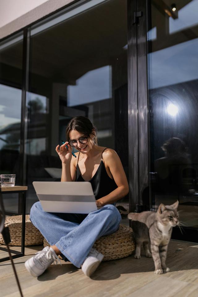 Woman sitting cross-legged on floor using laptop with a cat nearby, enjoying daylight indoors.
