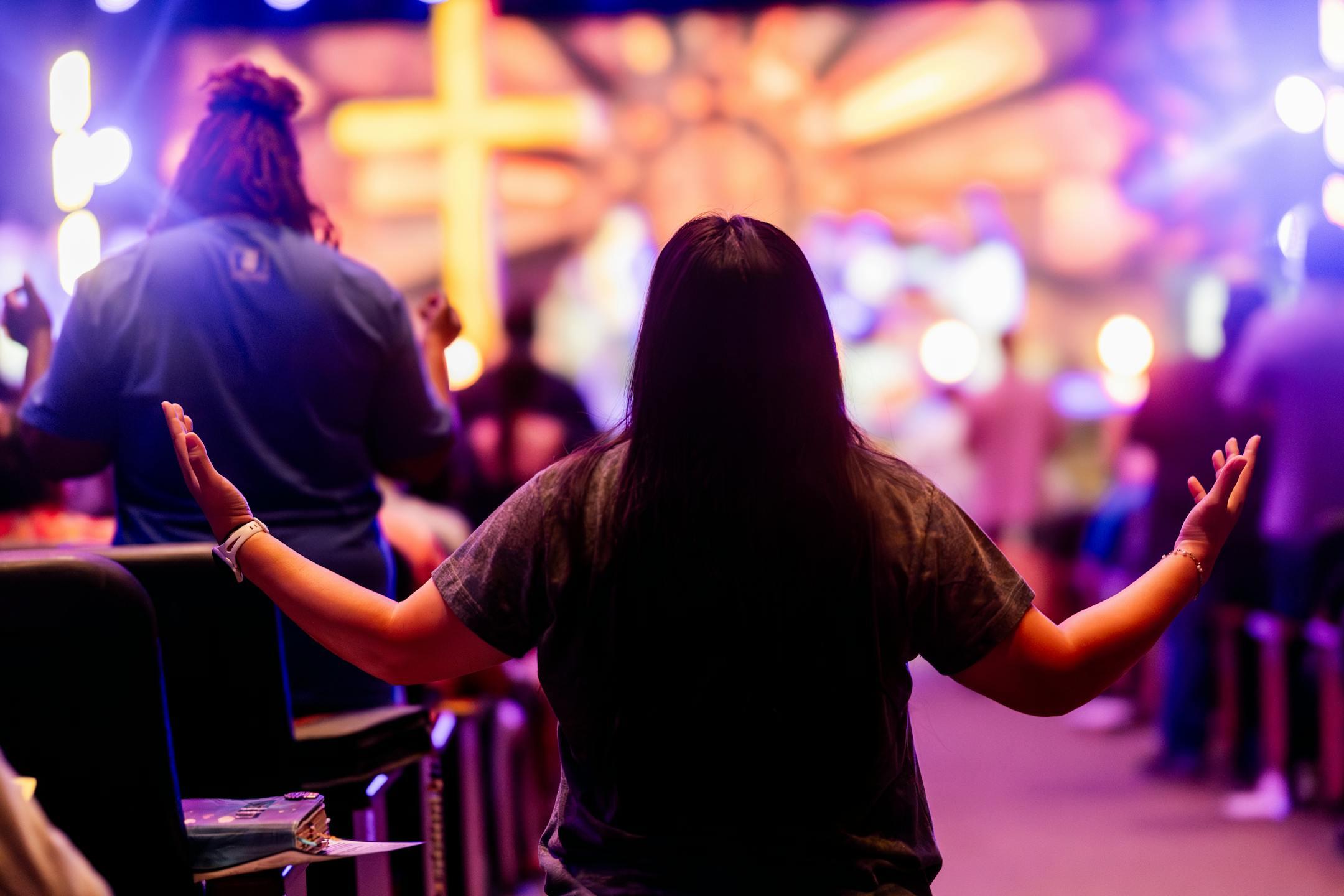 Back view of people engaged in a vibrant religious service with colorful lighting and a cross.