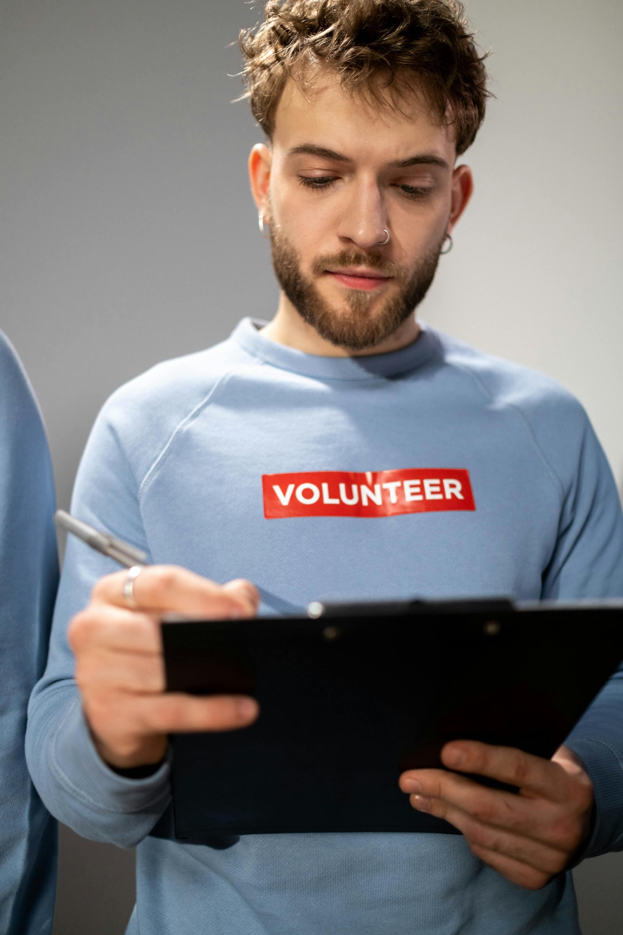Young adult male volunteer with beard writing on clipboard, wearing a volunteer shirt indoors.